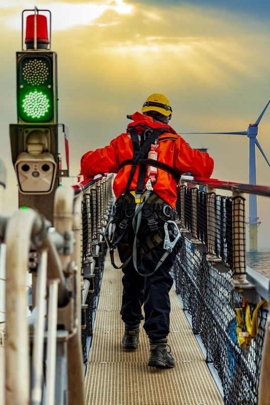 A wind turbine technician wearing safety gear and an orange jacket walking across a motion compensated gangway used for transferring personnel from a vessel to an offshore wind turbine.